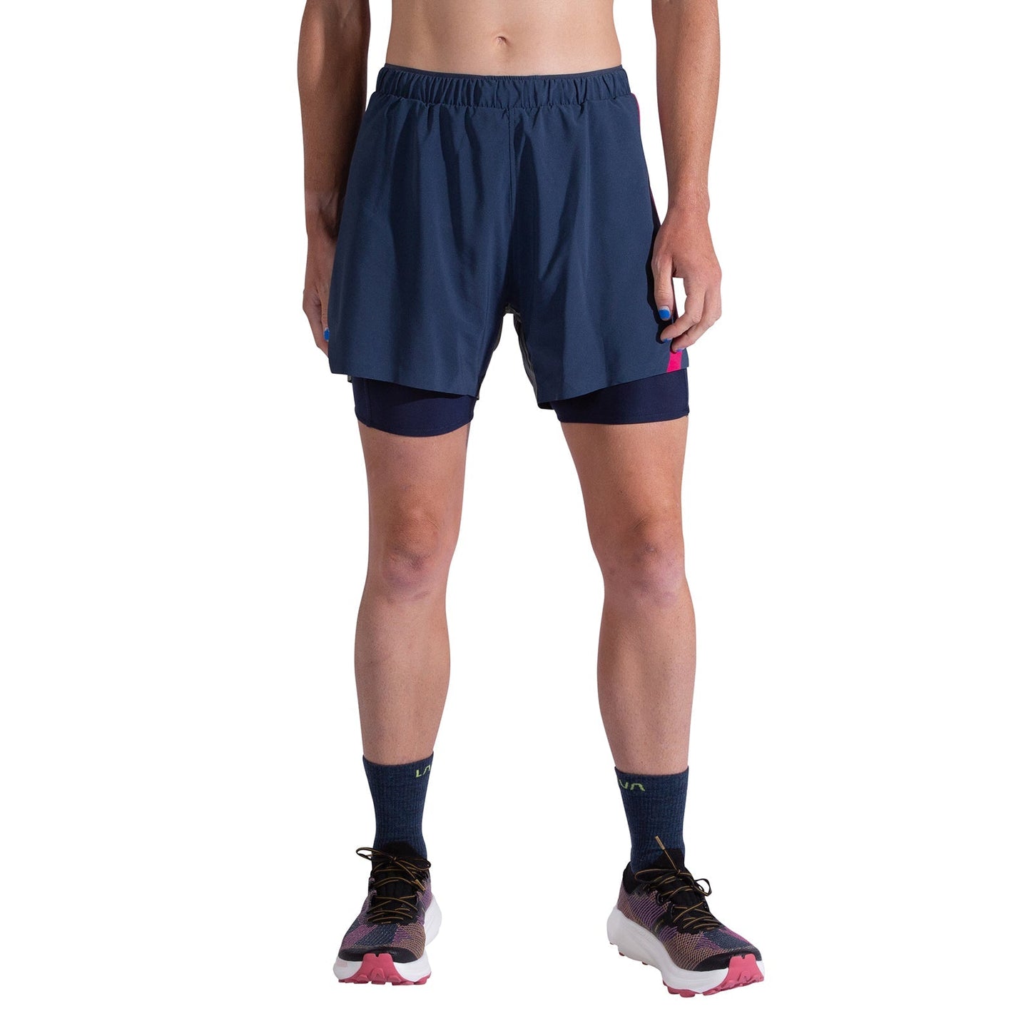 A person stands against a plain white background wearing La Sportiva Flow 5 Inch Shorts Women's in dark blue, navy socks, and athletic shoes with pink soles; the image is cropped from waist to feet.