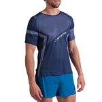 A man wears a La Sportiva Flow T-Shirt Men's in navy blue and gray with geometric patterns and light blue running shorts, standing against a white background.