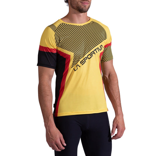 A man wears the La Sportiva Flow T-Shirt Men's—a yellow short-sleeve trail-running shirt with black and red accents—standing relaxed against a white background. This lightweight, breathable top is built for active performance.