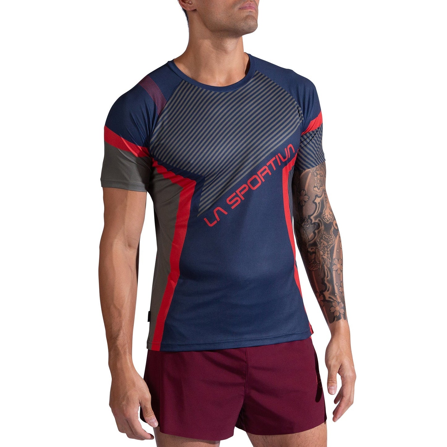 A man wears the La Sportiva Flow T-Shirt Men's, a navy and gray lightweight breathable top with red accents and "La Sportiva" branding, paired with burgundy shorts. He has a full sleeve tattoo on his left arm and stands before a white background.