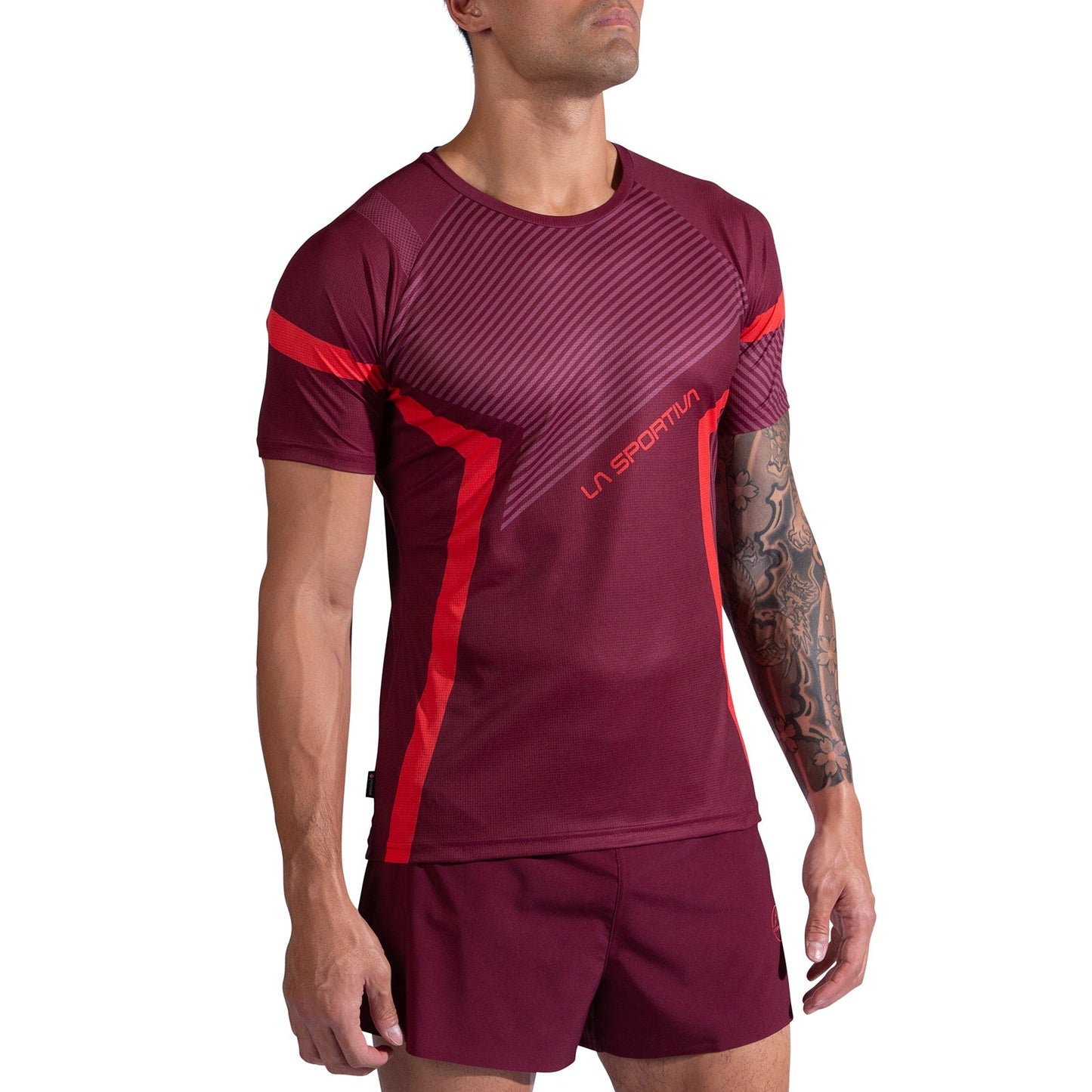 A man stands with his arms relaxed, wearing the La Sportiva Flow T-Shirt Men's in maroon with red accents and matching maroon shorts. A tattoo sleeve is visible on his right arm.