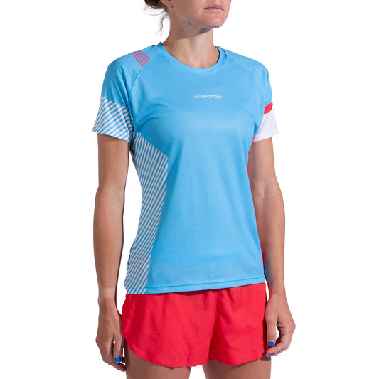 A woman stands against a white background wearing the La Sportiva Flow T-Shirt Women's in light blue with white and red accents, paired with red shorts. The sporty, short-sleeved shirt is perfect for athletes and her face is partially out of frame.