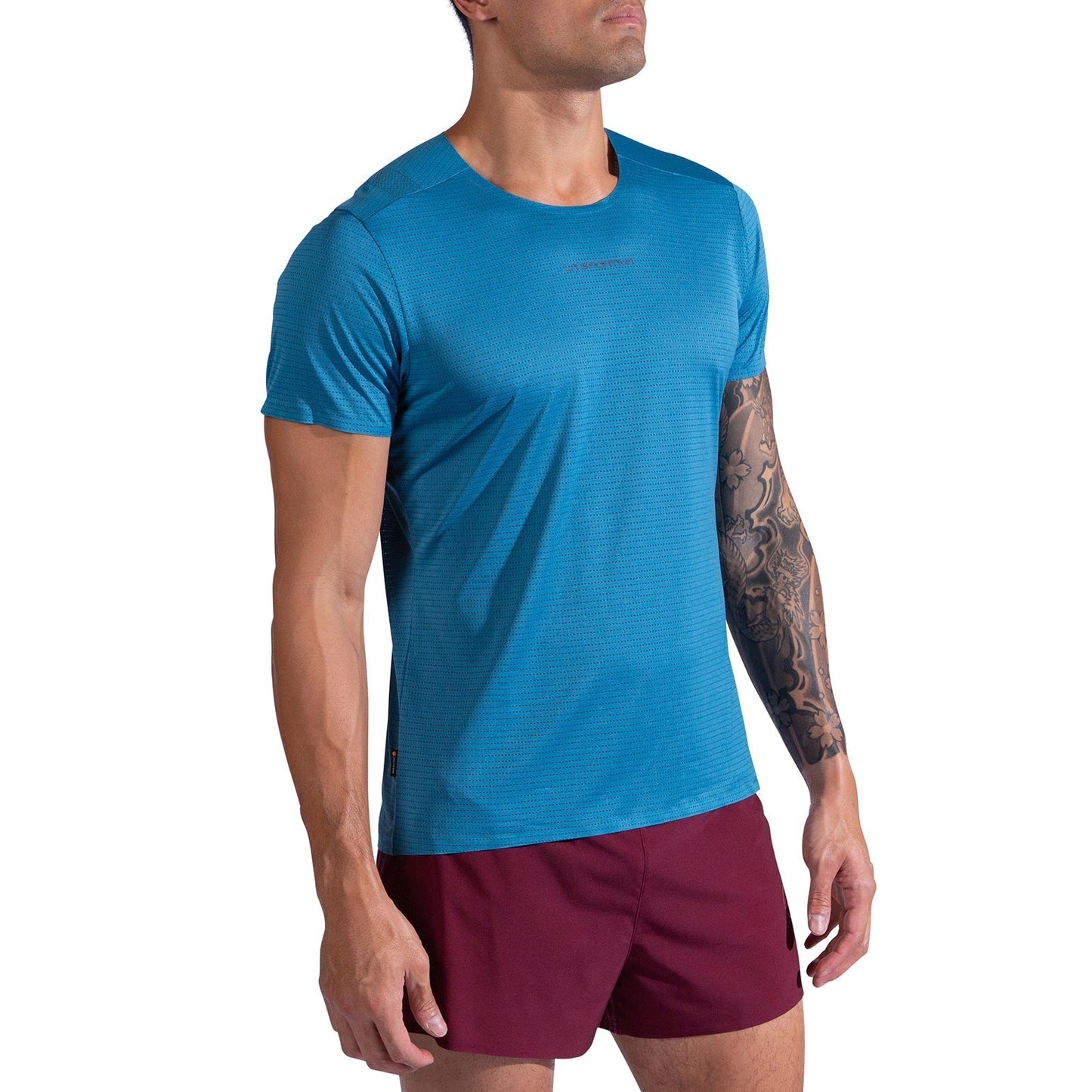 A person with a tattoo sleeve on their left arm stands against a white background, wearing the La Sportiva Pure T-Shirt Men's in blue and maroon shorts. The short-sleeve running tee is made from recycled polyester.