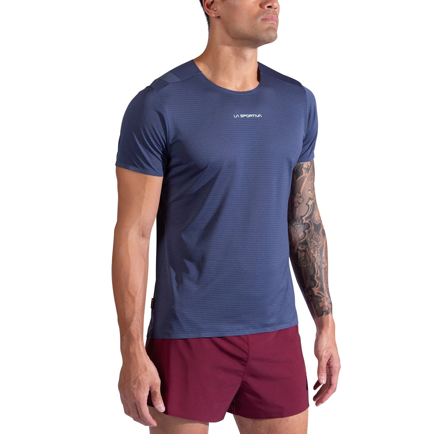 A man wearing the La Sportiva Pure T-Shirt Men’s in blue and maroon shorts stands against a white background. The short-sleeve running tee is made from recycled polyester. He has a tattoo sleeve on his left arm and looks slightly upwards.