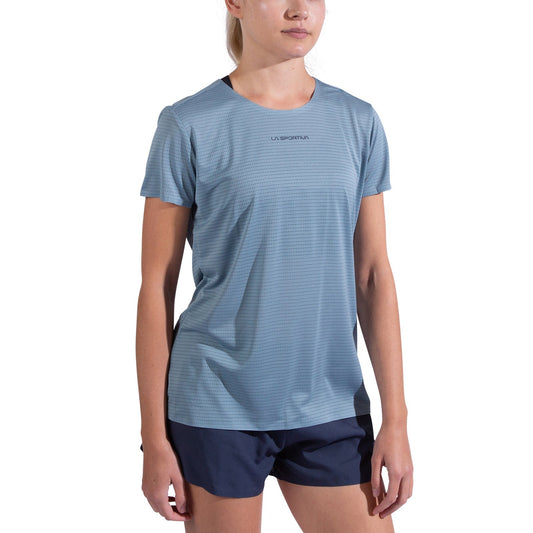 A woman wears the La Sportiva Pure T-Shirt Women's in light blue, made from recycled polyester, with navy blue shorts, standing against a plain white background. The shirt displays "La Sportiva" on the chest.