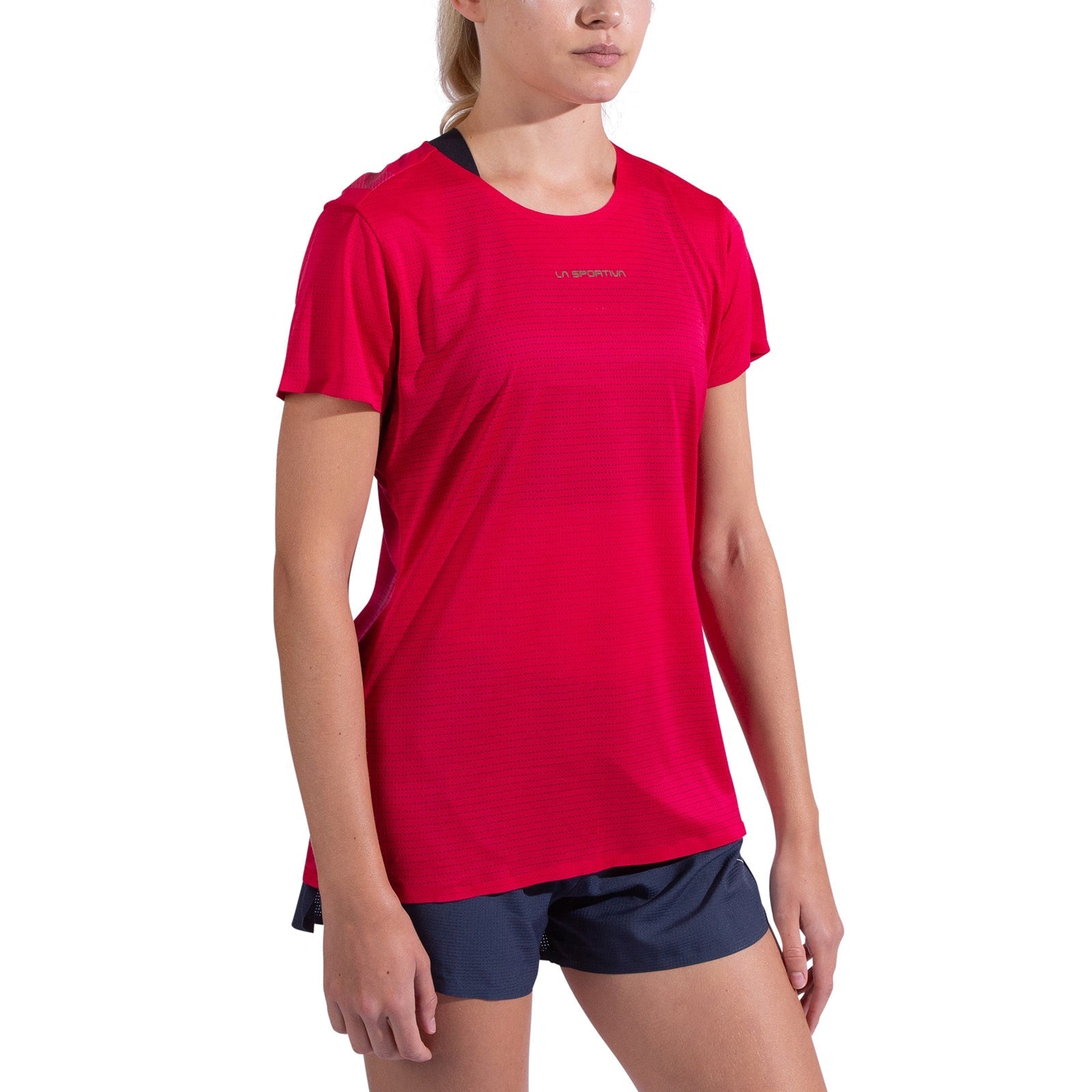 A woman stands against a white background, wearing the La Sportiva Pure T-Shirt Women's in bright pink with short sleeves and navy blue shorts, her arms relaxed by her sides as she faces slightly left.