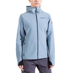 A woman wearing the La Sportiva Descender Storm Jacket—light blue softshell with cozy fleece interior, front zipper, thumbholes, and La Sportiva logo on chest and sleeve—pairs it with dark gray pants against a white background.