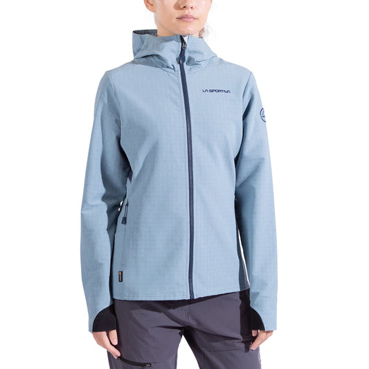 A woman wearing the La Sportiva Descender Storm Jacket—light blue softshell with cozy fleece interior, front zipper, thumbholes, and La Sportiva logo on chest and sleeve—pairs it with dark gray pants against a white background.
