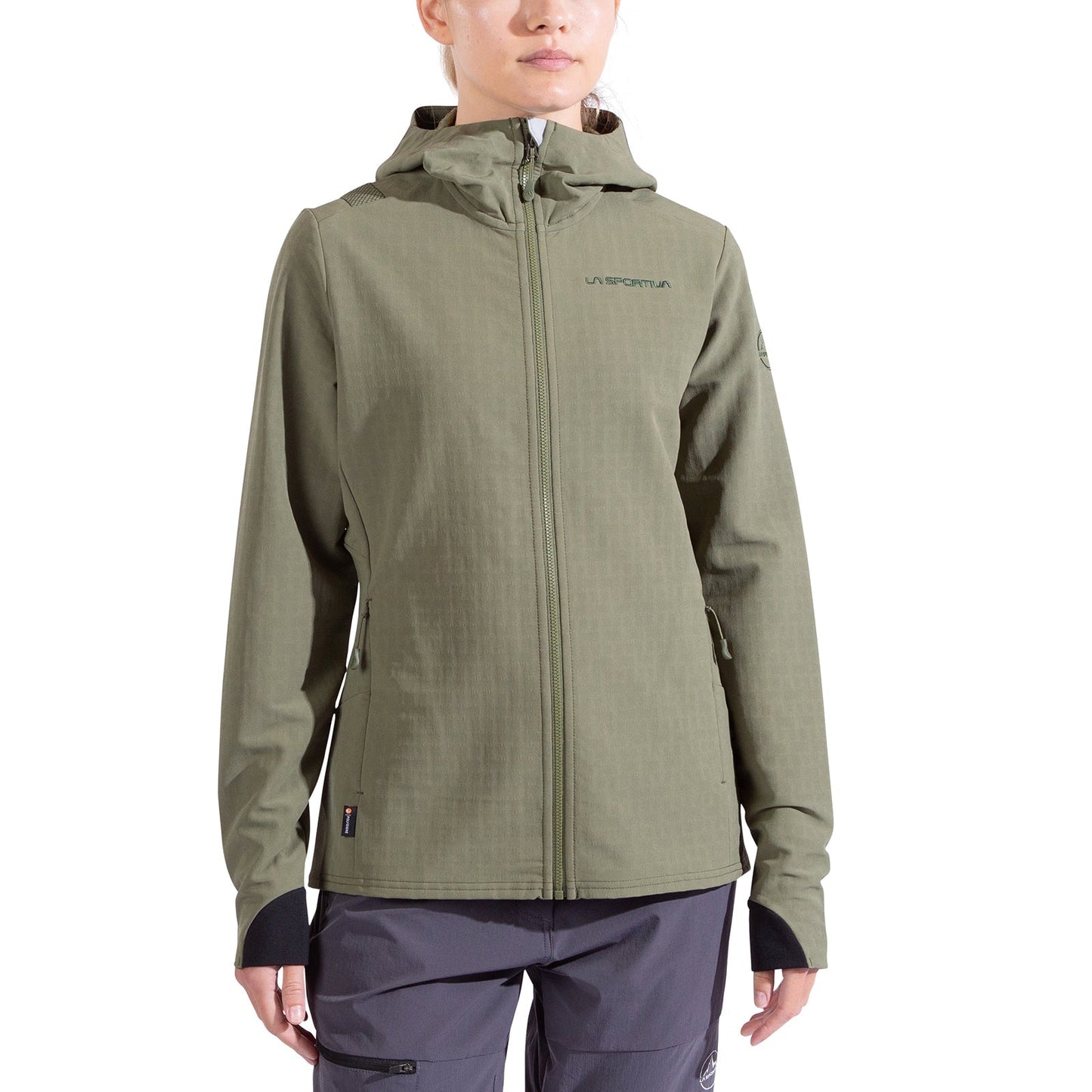 A person wears the La Sportiva Descender Storm Jacket Women's in light olive green, featuring a fleece interior, black cuffs, and a subtle chest logo, paired with gray pants and standing against a white background.