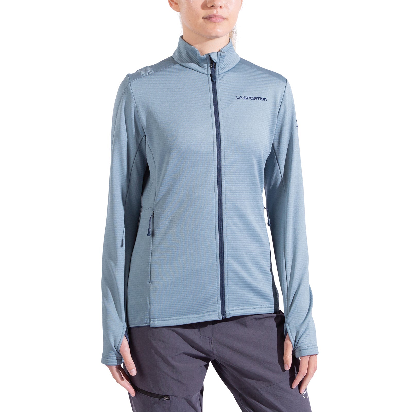 A woman wearing a light blue La Sportiva Chill Thermal Jacket with long sleeves, side pockets, and chest logo pairs it with gray pants while standing against a white background.