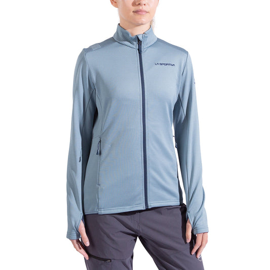 A woman wearing a light blue La Sportiva Chill Thermal Jacket with long sleeves, side pockets, and chest logo pairs it with gray pants while standing against a white background.