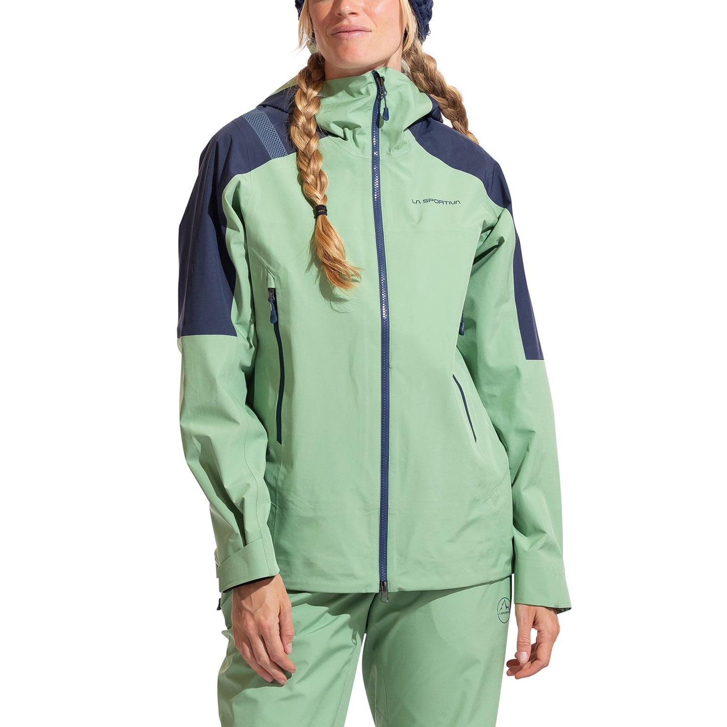 A woman in the La Sportiva Crest Evo Shell Jacket Women's, light green and dark blue with matching pants, stands facing forward. She wears braids and a knit hat. The jacket includes a helmet-compatible hood for versatility.