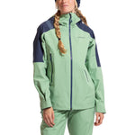 A woman in the La Sportiva Crest Evo Shell Jacket Women's, light green and dark blue with matching pants, stands facing forward. She wears braids and a knit hat. The jacket includes a helmet-compatible hood for versatility.