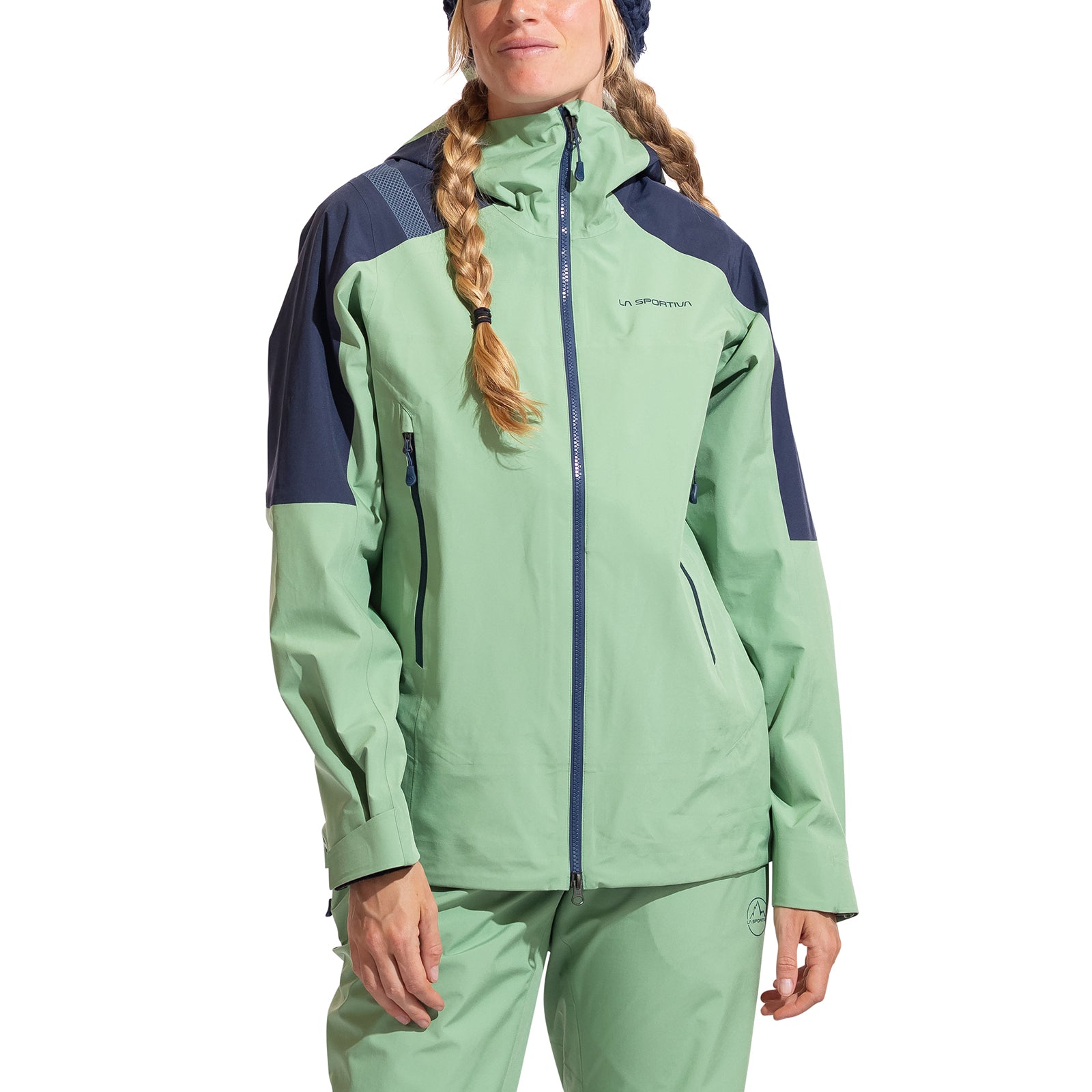 A woman in the La Sportiva Crest Evo Shell Jacket Women's, light green and dark blue with matching pants, stands facing forward. She wears braids and a knit hat. The jacket includes a helmet-compatible hood for versatility.