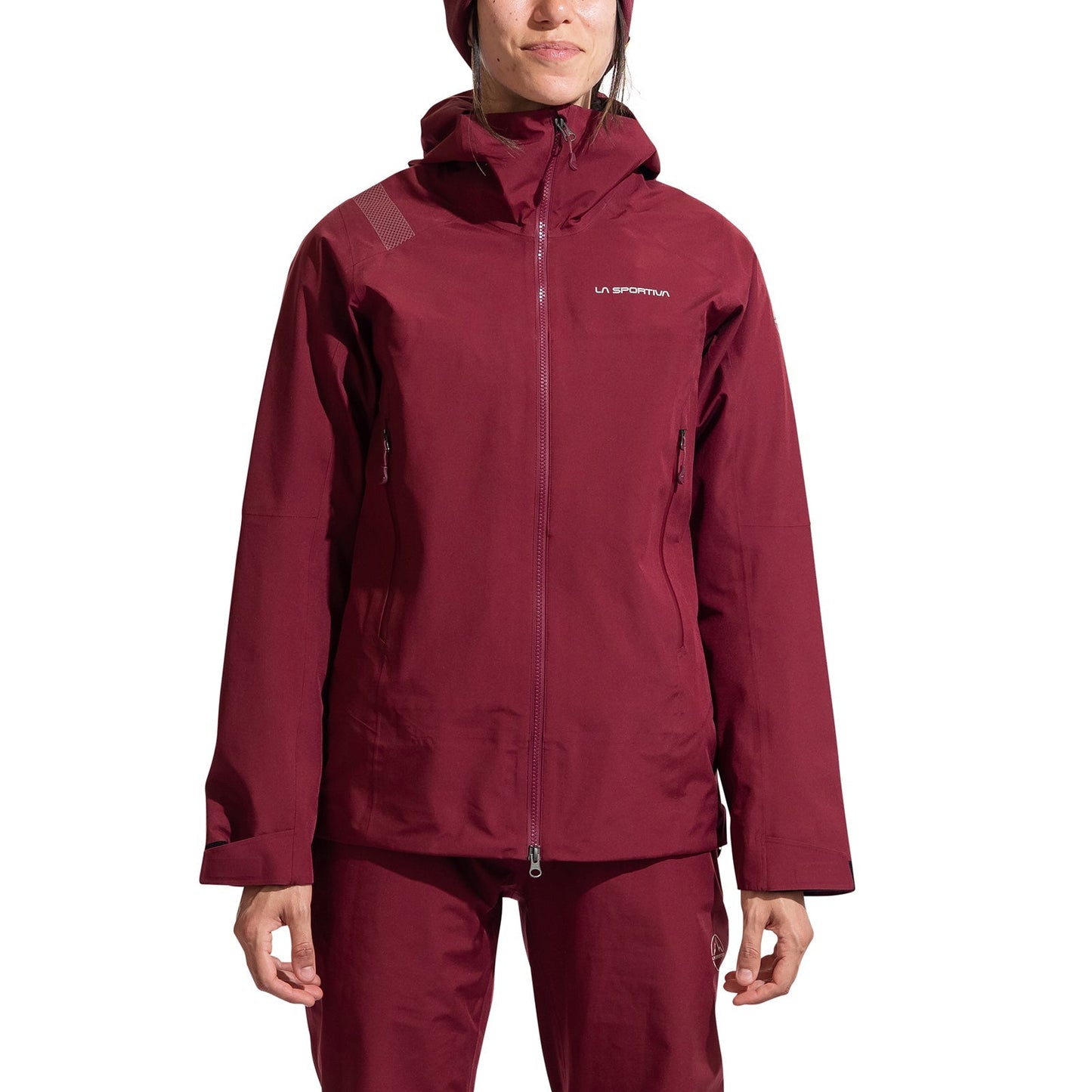 A woman models the La Sportiva Crest Evo Shell Jacket, featuring a maroon color, front zipper, two side pockets, chest logo, helmet-compatible hood, and matching pants, standing against a white background.