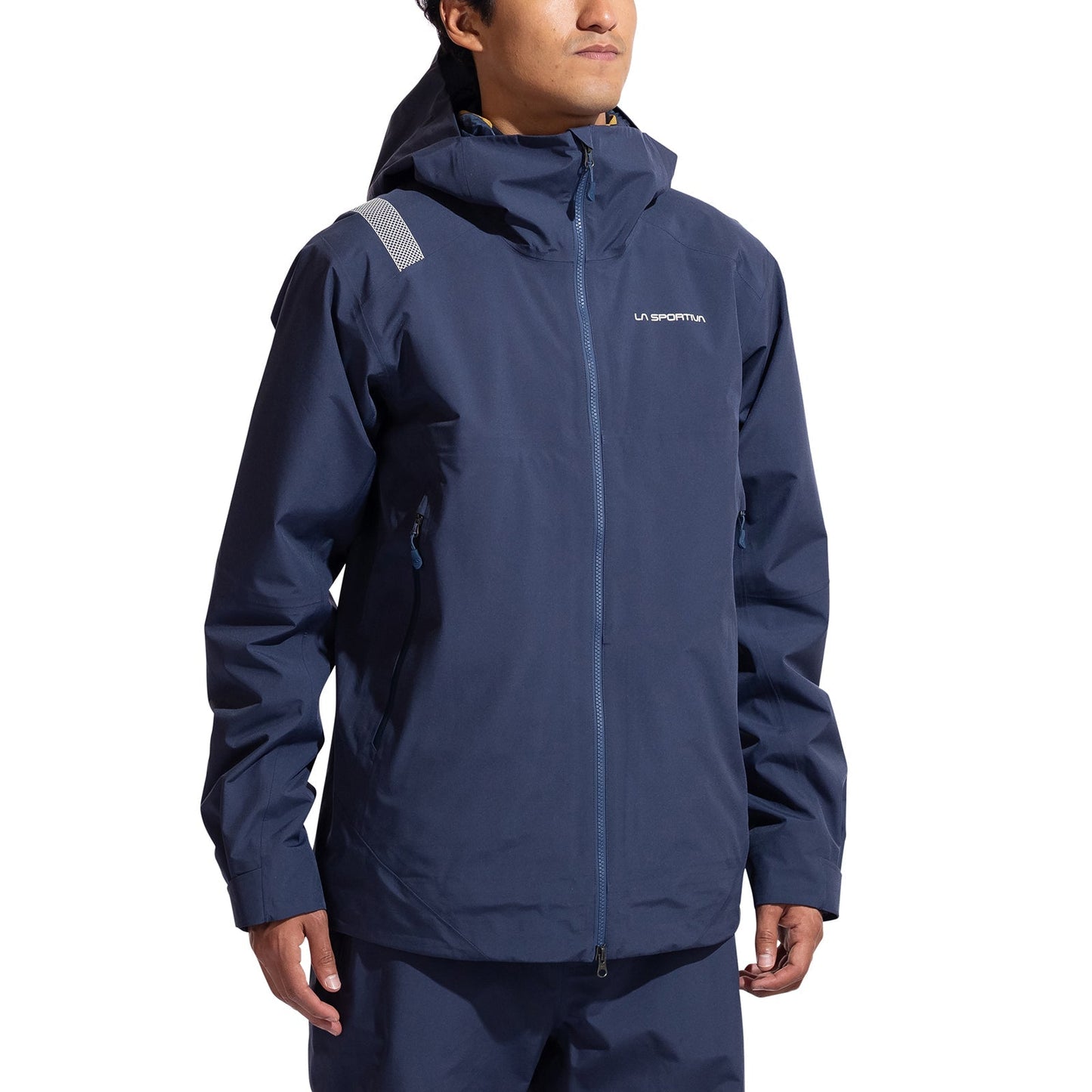 A person in a La Sportiva Crest Evo Shell Jacket Men's, navy blue with zippered pockets, helmet-compatible hood, and a reflective patch on the left shoulder, stands against a plain white background.