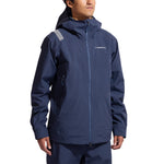 A person in a La Sportiva Crest Evo Shell Jacket Men's, navy blue with zippered pockets, helmet-compatible hood, and a reflective patch on the left shoulder, stands against a plain white background.