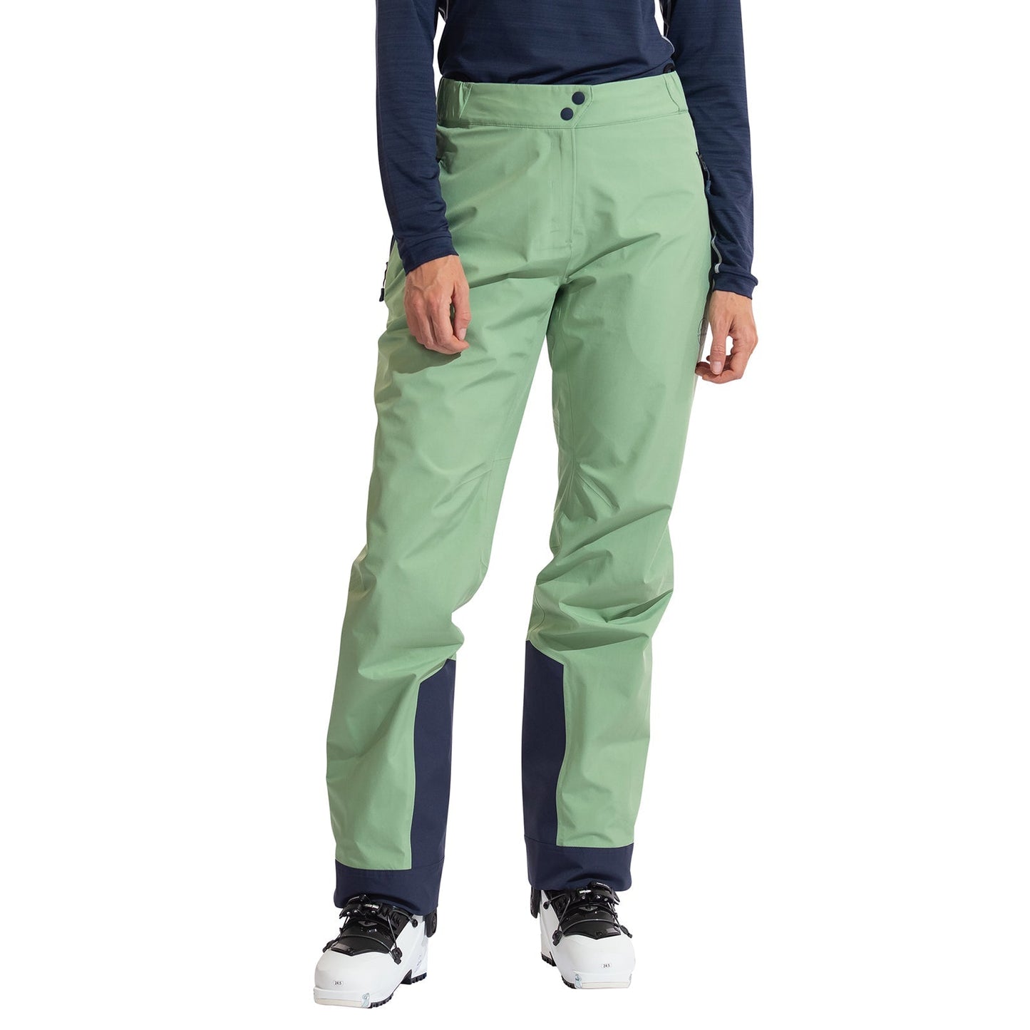A woman stands against a white background wearing La Sportiva Crest Evo Shell Pants Women's in light green with dark navy ankle accents, paired with a navy long-sleeve top and white ski boots.