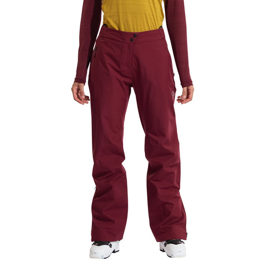 A person wearing La Sportiva Crest Evo Shell Pants Women's in burgundy, a yellow shirt, and a long-sleeve burgundy top stands with legs apart in white ski boots.