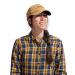 A person with long brown hair in a side braid wears the La Sportiva Setter Cap and a yellow, blue, and brown plaid shirt, smiling and looking slightly to the side against a white background.