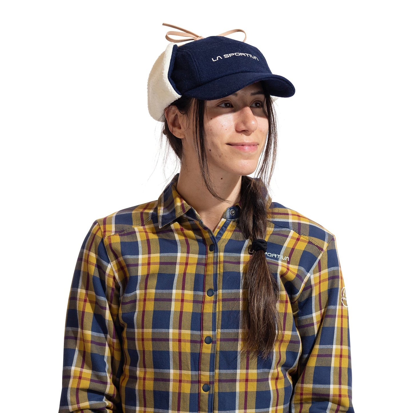 A woman in a navy La Sportiva Forest Earflap Cap and a yellow, blue, and brown plaid button-up shirt smiles slightly to the side against a white background.