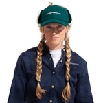 A woman with long blonde braids wears a green La Sportiva Forest Earflap Cap with sherpa lining, a navy plaid shirt, and a white undershirt, standing against a white background.