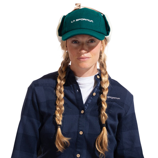 A woman with long blonde braids wears a green La Sportiva Forest Earflap Cap with sherpa lining, a navy plaid shirt, and a white undershirt, standing against a white background.