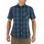 A person wearing the La Sportiva Nomad Short Sleeve Shirt Men's—a blue plaid, recycled polyester button-up with a chest pocket—paired with khaki shorts, stands against a plain white background.