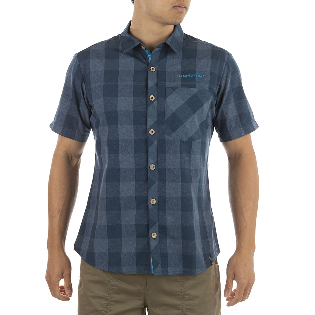 A person wearing the La Sportiva Nomad Short Sleeve Shirt Men's—a blue plaid, recycled polyester button-up with a chest pocket—paired with khaki shorts, stands against a plain white background.