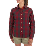 A woman wears the La Sportiva Andes Long Sleeve Shirt Women's in red and dark gray plaid, paired with khaki shorts. The shirt, made from recycled polyester, features visible La Sportiva branding on the chest and sleeve.