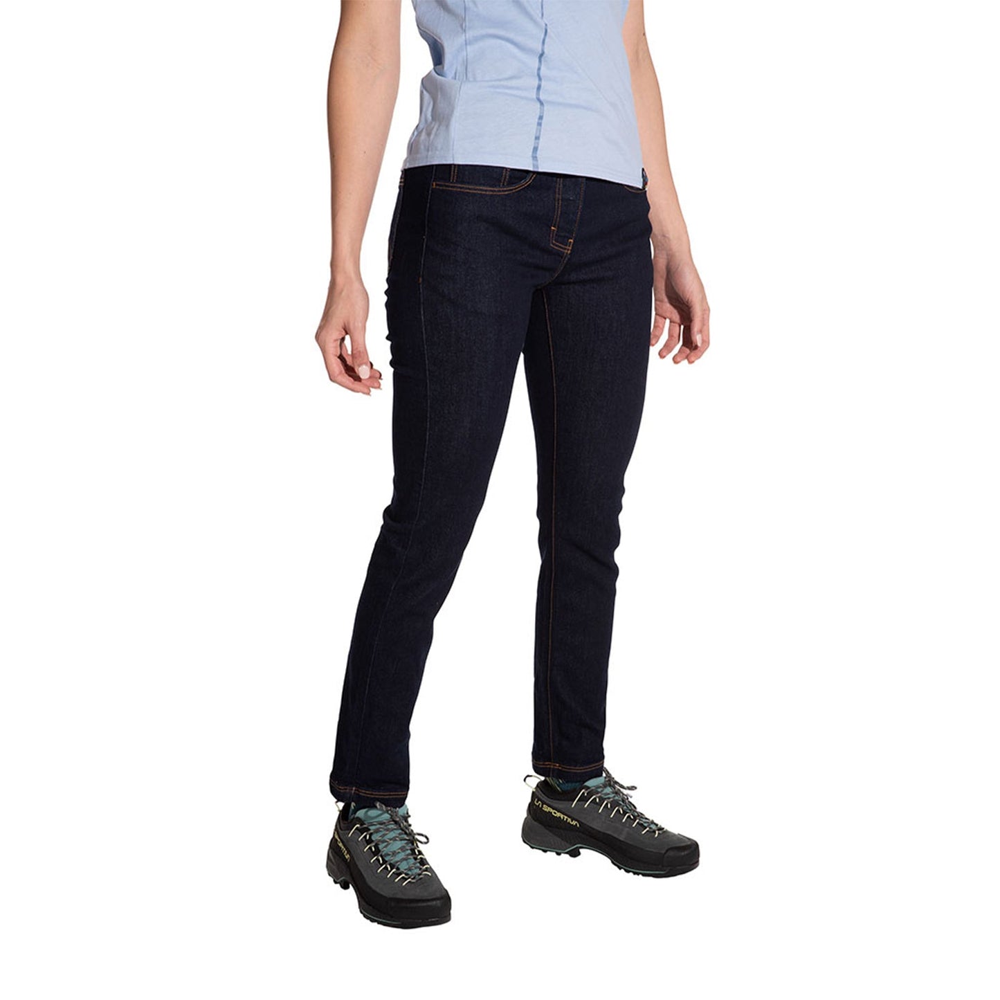 A woman wearing La Sportiva Eldo Jeans 2.0 and a light blue shirt stands against a white background, shown from shoulders to feet. She also wears black hiking shoes.