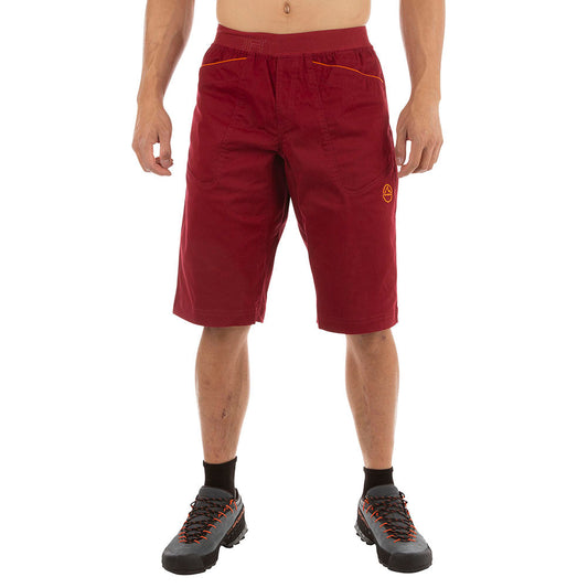 A person wears La Sportiva Flatanger Shorts Men's (2023) in red-orange, with black socks and black trail shoes with orange laces, standing against a white background. The cotton-spandex blend offers comfort and flexibility. The person's head is not visible.