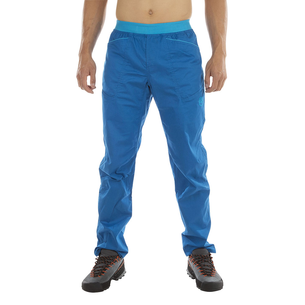 A person wearing La Sportiva Roots Pants Men's (2024) in bright blue and hiking boots stands facing forward against a white background, with only their lower body visible.