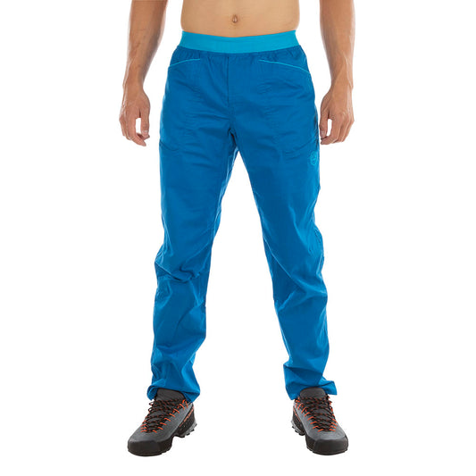 A person wearing La Sportiva Roots Pants Men's (2024) in bright blue and hiking boots stands facing forward against a white background, with only their lower body visible.