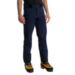 A person wearing La Sportiva Excelsior Pants Men's in navy blue with black accents and yellow hiking boots, designed for mountaineering, stands against a white background. Only the lower half of the body is visible.
