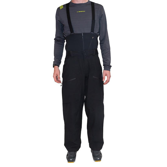 A person wearing a long-sleeve gray shirt, gray shoes, and La Sportiva Chaser EVO Shell Bib Pants Men's in black with suspenders, standing facing forward against a white background.