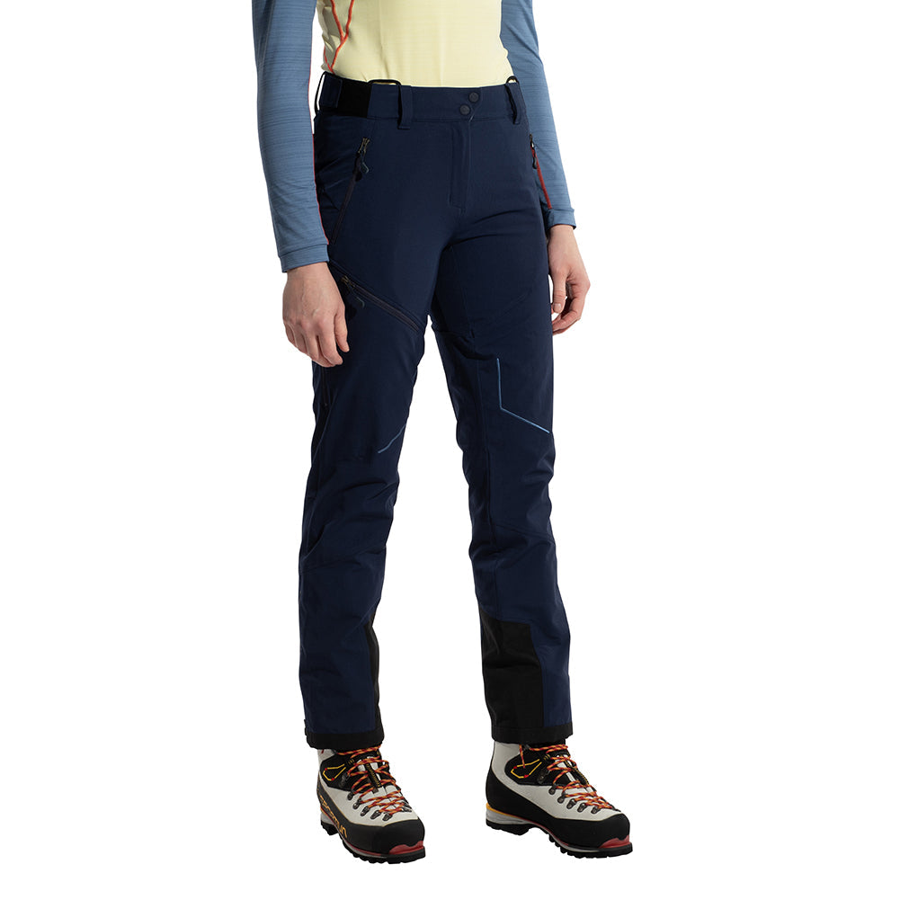 A woman stands against a white background wearing La Sportiva Excelsior Pants, a yellow and blue long-sleeve shirt, and mountaineering boots with orange laces.