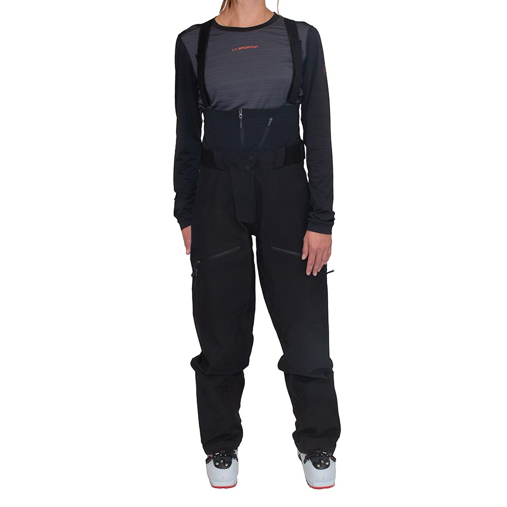 A woman models the La Sportiva Chaser EVO Shell Bib Pants with a high waist, paired with a long-sleeve black top and ski boots. She stands facing forward against a white background, arms relaxed and ready for snow adventures.