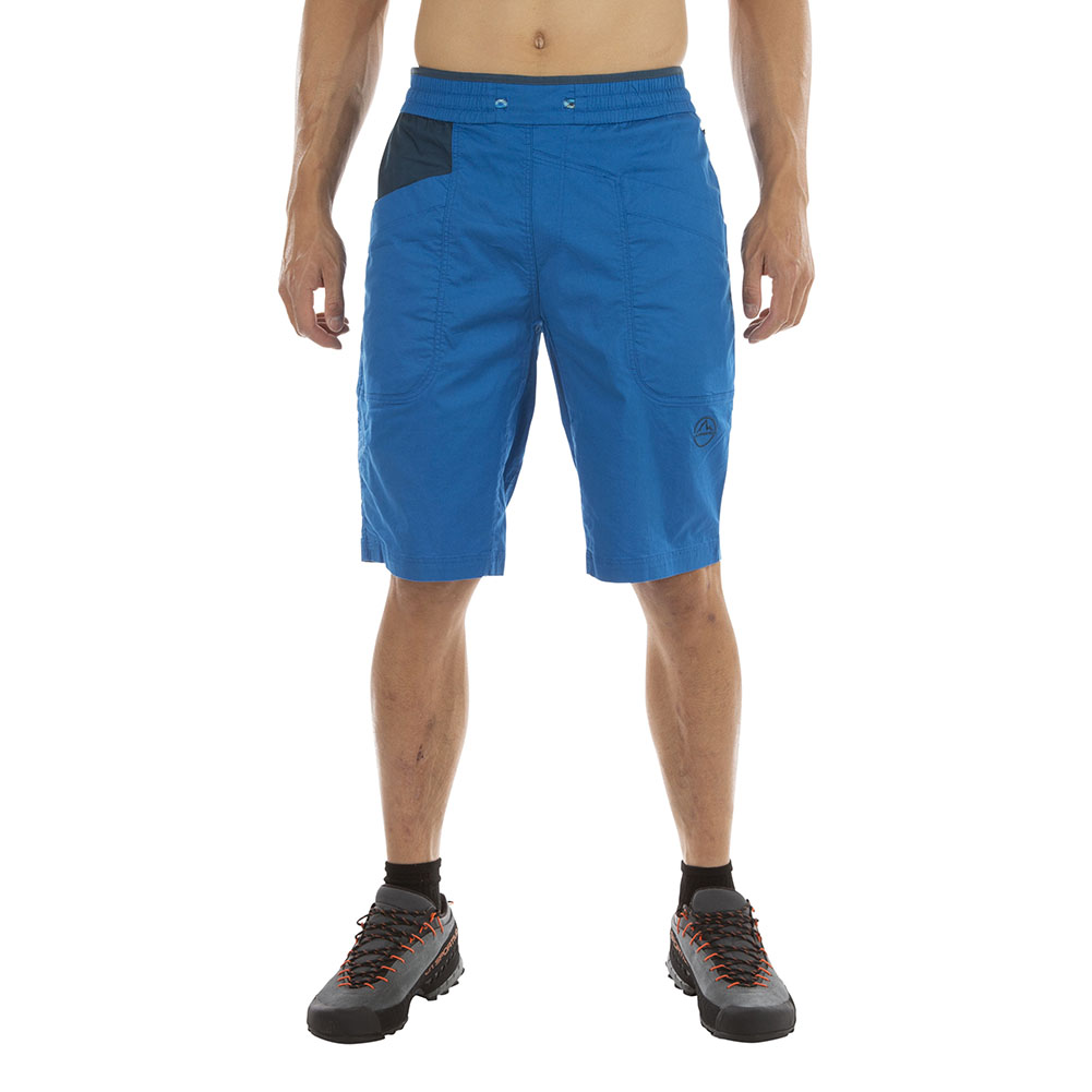A person wears La Sportiva Bleauser Shorts Men's—bright blue, organic cotton with gusseted crotch and pockets—and black hiking shoes, standing against a white background. Only their body from neck to feet is visible.