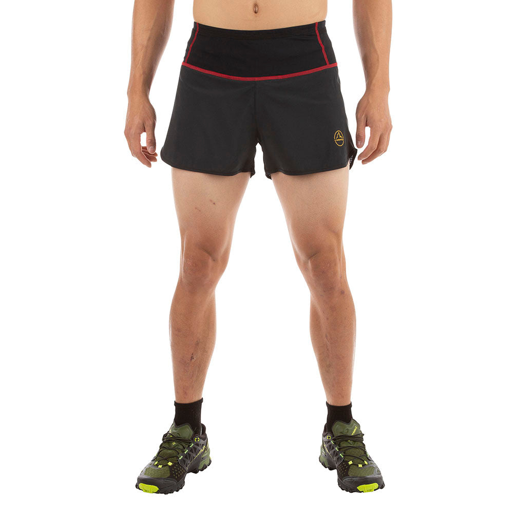 A person wears La Sportiva Tempo Shorts Men's—black with red trim and logo, featuring a lightweight recycled-material waistband—paired with black socks and green running shoes. Only the lower body is shown against a white background.